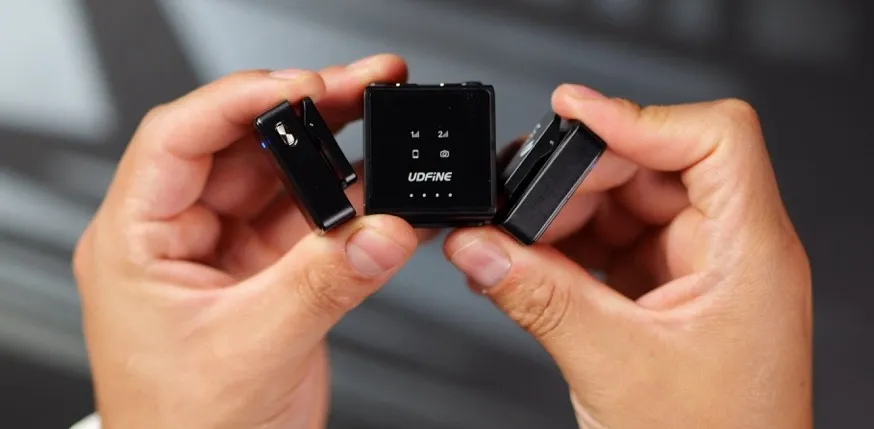 Udfine Mic Lite en uso durante vlogging outdoors, transmisor clip-on en camisa, receptor conectado a smartphone Samsung, con caja de carga abierta mostrando 33h total, fondo con paisaje ecuatoriano y medición de rango 80m visible.